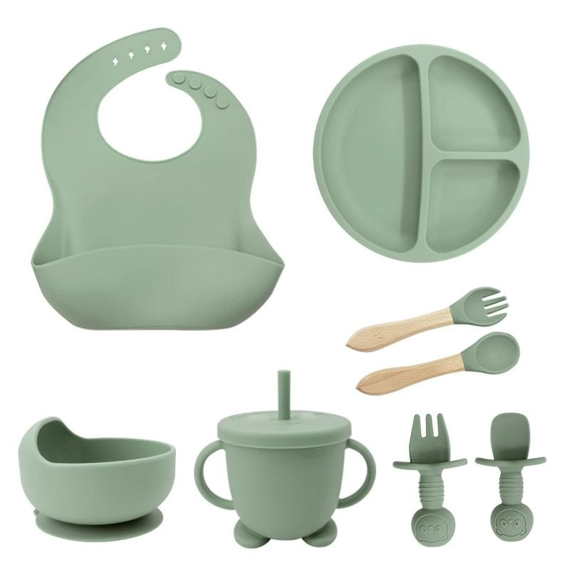 Set of green silicone baby utensils including a bib, plate, cup, and bowls on a white background.