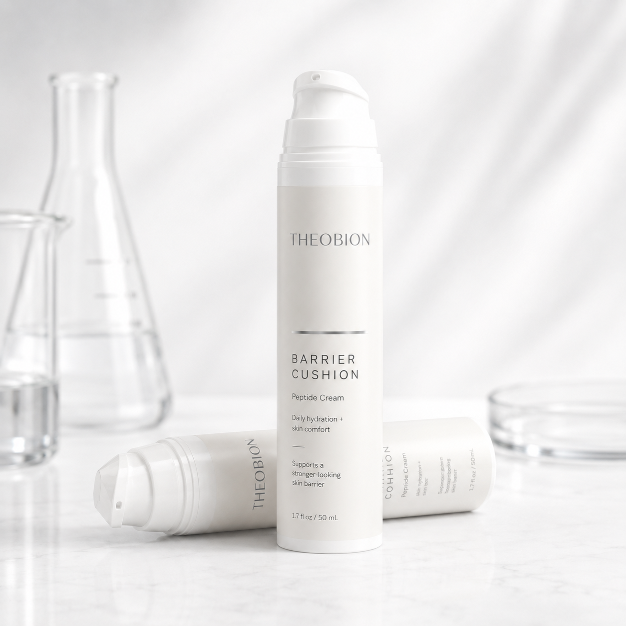 A white pump bottle of THEOBION Barrier Cushion Peptide Cream stands upright on a light marble surface, with two additional bottles lying horizontally in the foreground and blurred glassware in the background.